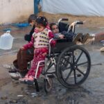 Palestinian children sit in a wheelchair while they make their