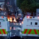 People walk near official vehicles as flames rise during a