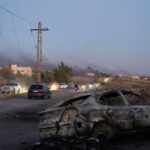 Smoke rises as a damaged car remains along a street,