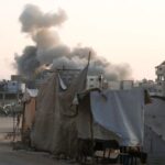 Palestinians stand as smoke rises following an Israeli strike, in