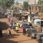 A cyclist and motorcyclists past cars parked on the roadside,